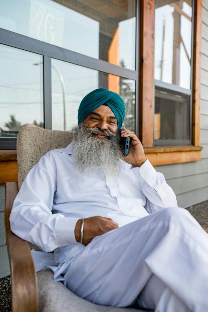 Adult Sikh man wearing traditional clothing, sitting outdoors, talking on smartphone.